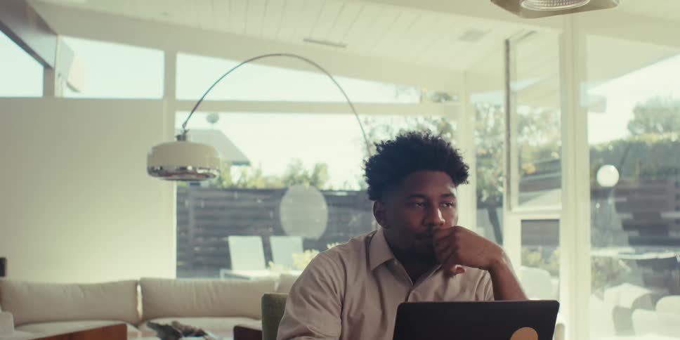 A person is sitting at a table working on a laptop in a bright modern white room with large windows overlooking a lush garden