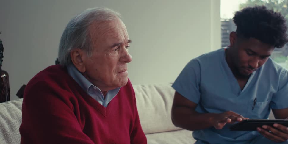An elderly man in a red sweater sits next to a healthcare worker in blue scrubs who is using a tablet