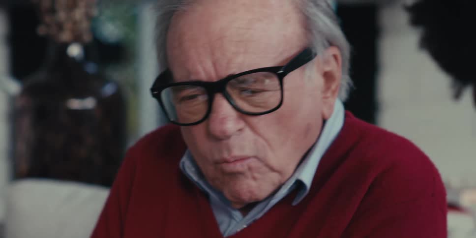 An elderly man in a red sweater and glasses talking to a healthcare worker in blue scrubs