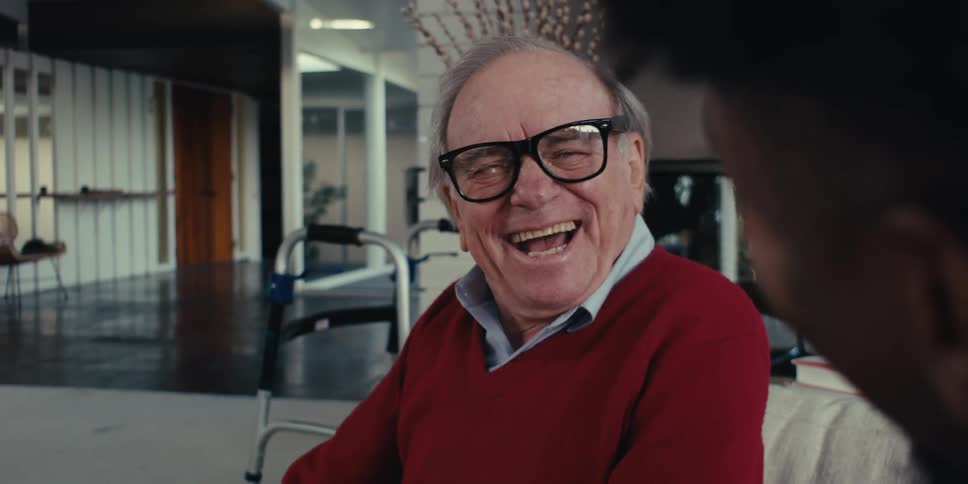 Elderly man sitting on a couch laughs while talking with a medical professional