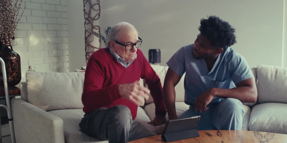 A home health aide talks to an elderly man while they look at an electronic tablet