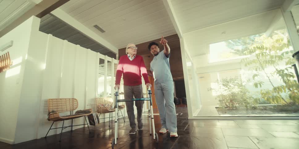 Elderly man walking with the help of a walker next to a nurse in the hallway of a house