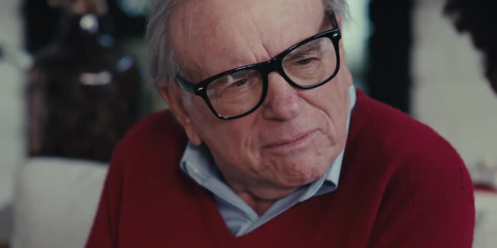 Closeup of an elderly man wearing glasses talking to another person on the couch in a modern spacious living room