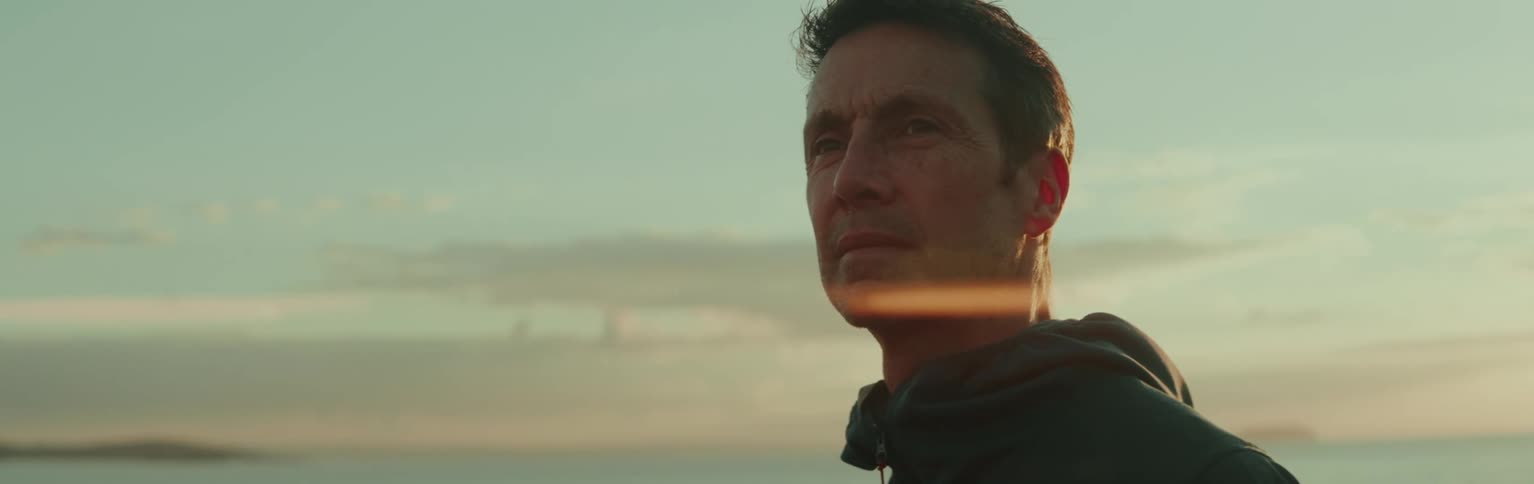 A person in a hoodie stands near a calm sea during sunset with a serene sky and soft clouds in the background