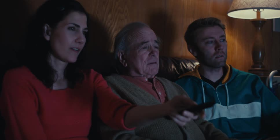 Elderly man sitting in center of two young adults watching television in living room at night