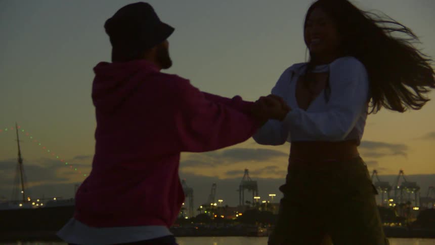 Two people on roller skates are dancing holding hands near a waterfront at sunset with an industrial skyline in the background