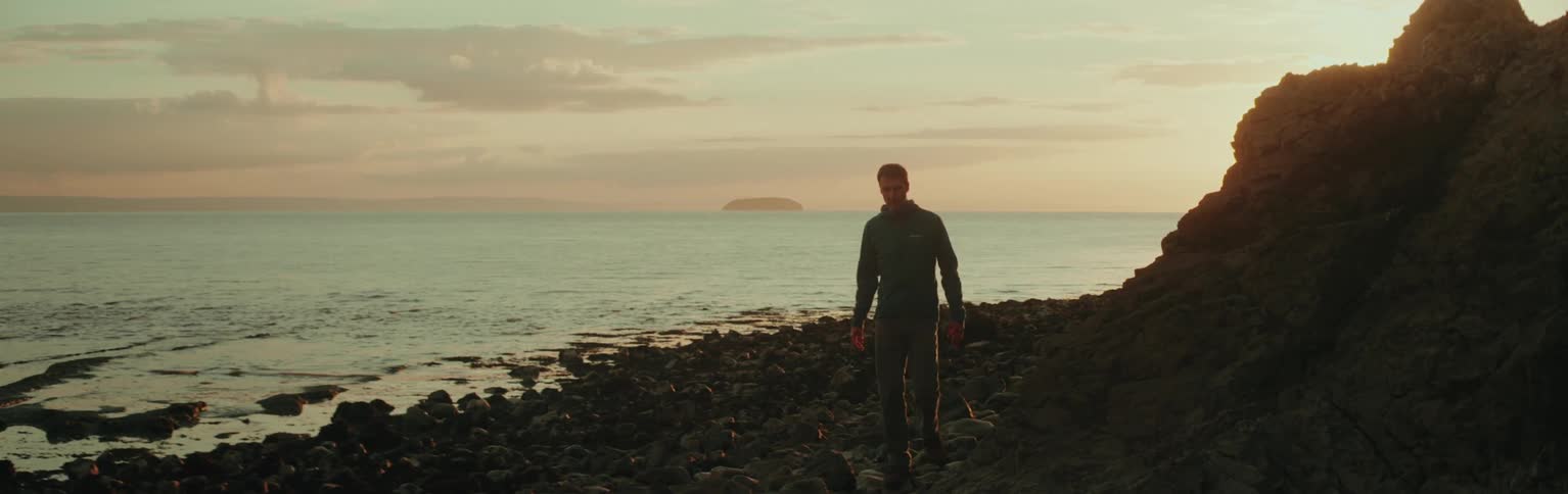 A man walks along a rocky ocean coastline at sunset