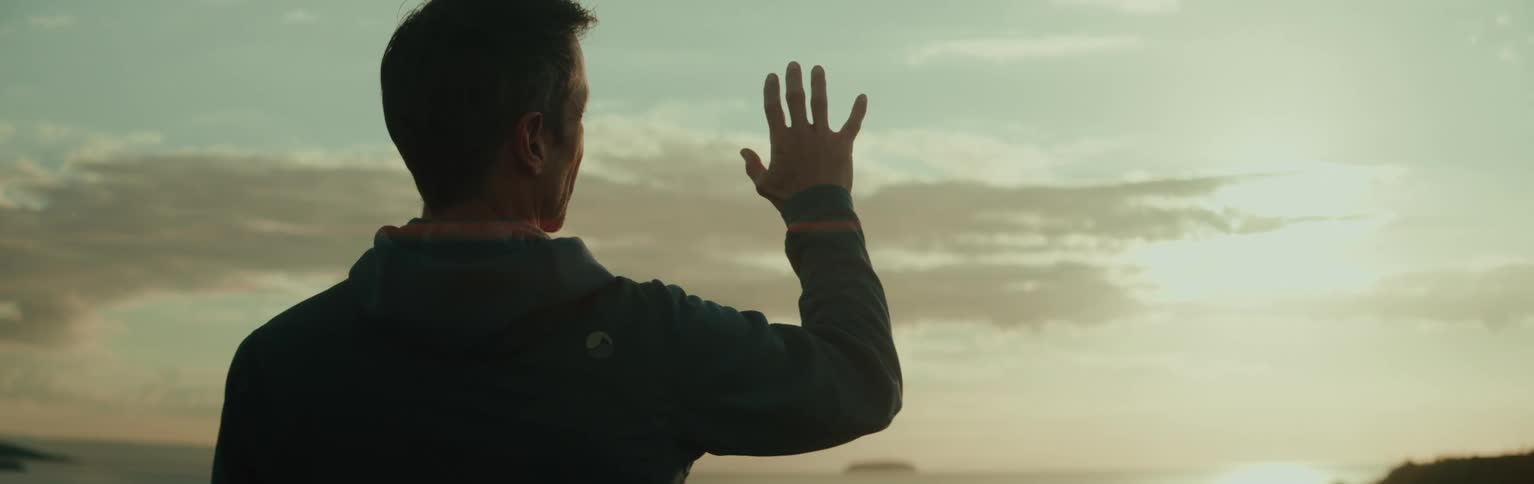 A person is standing outdoors facing a sunset or sunrise with an arm raised and fingers spread against a backdrop of clouds and a bright sky