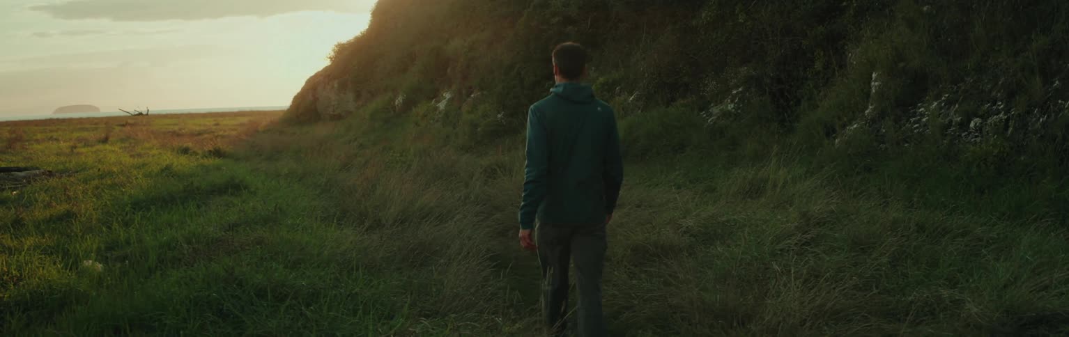 A person in a green hoodie is walking through a grassy field towards a sunlit hillside