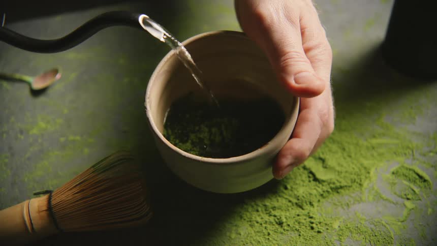 A hand is holding a bowl while hot water is being poured into it to prepare matcha tea