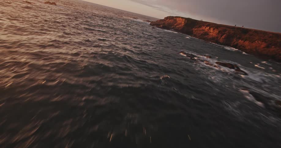 Roaring waves crash against the dark rocks at sunset dyeing the coastal landscape with golden hues and dramatic shadows