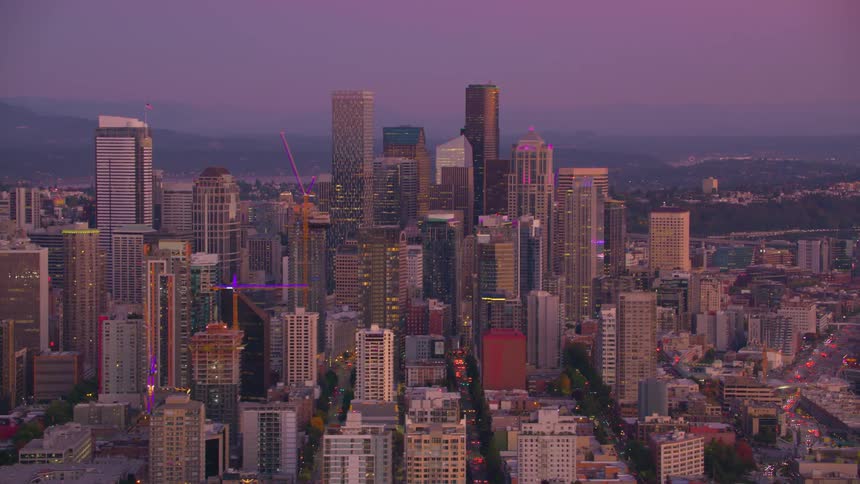 A sprawling cityscape glows with warm lights under a vibrant twilight sky featuring a cluster of tall buildings at its center