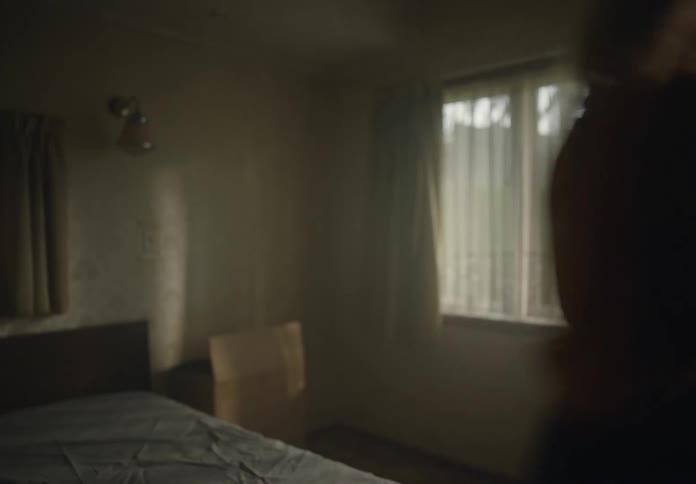 A woman is sitting on a bed then gets up and carries a cardboard box out of the room