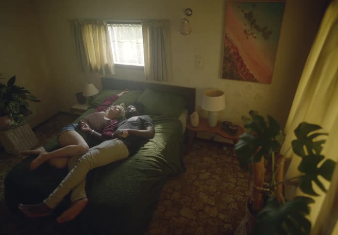 A couple is lying on a green bed in a dimly lit room with a window cuddling playfully