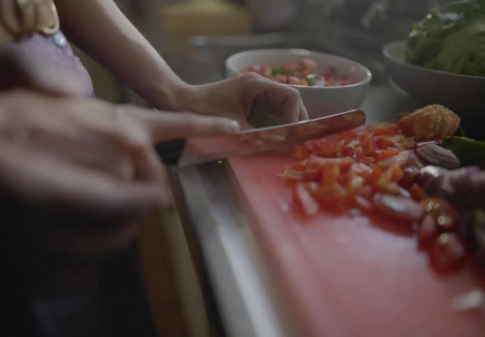 A person is slicing red bell peppers on a red cutting board with other chopped vegetables like onions and tomatoes visible nearby