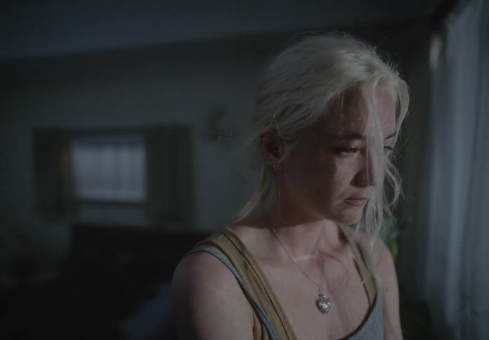 A young sad blonde woman stands in a dimly lit room staring at a window illuminated by the lights of a passing vehicle