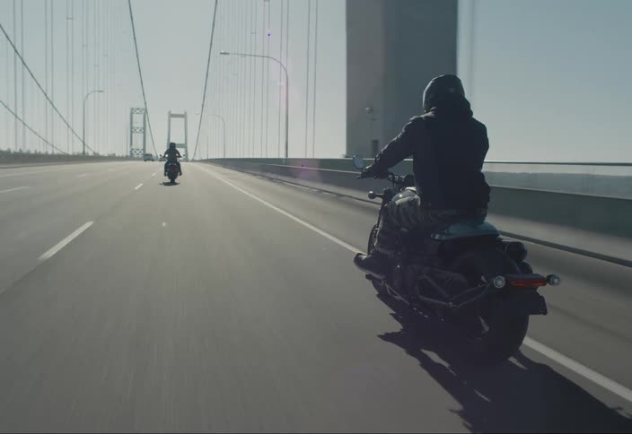 Motorcyclists ride across a bridge toward a forested shore during the day