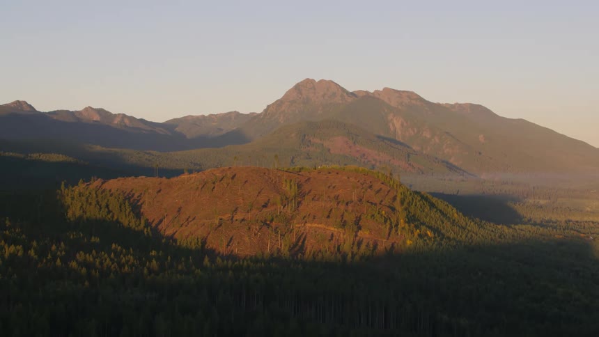 A rugged mountain peak partially illuminated by the warm glow of the setting sun rises above a densely forested valley