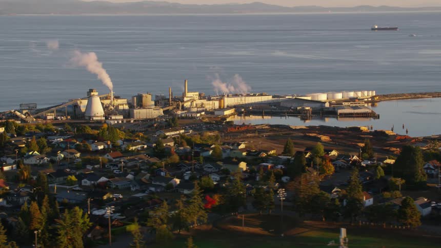 A sprawling industrial complex with smoking smokestacks sits along a harbor bordered by a densely populated town and a distant ocean horizon