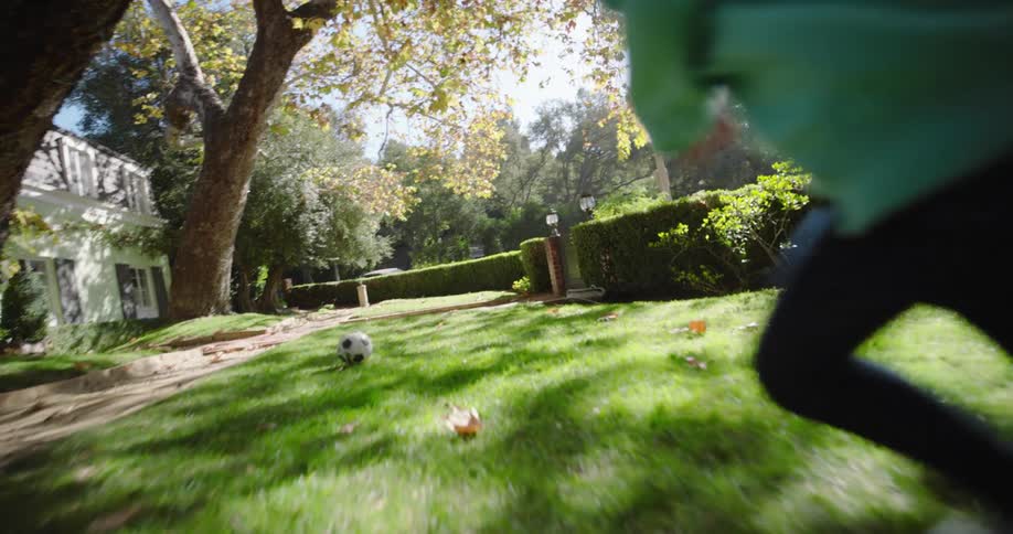 A child in a teal hoodie is running after a soccer ball on a grassy lawn with a large tree and a house in the background