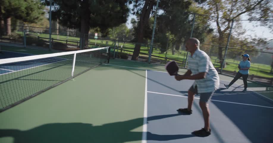 Two couples playing pickleball on an outdoor court surrounded by tall trees and a fence