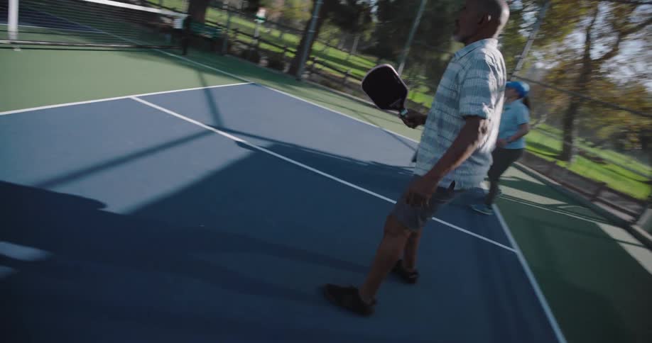 Two couples playing pickleball on an outdoor court surrounded by tall trees and a fence