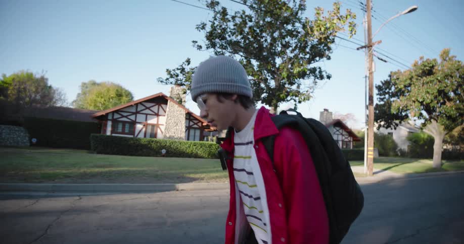 A teenager wearing a red jacket and black pants is skateboarding on a residential street at morning