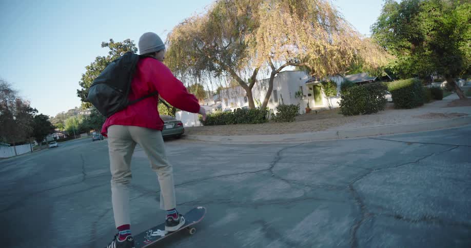 A young skateboarder wearing a red jacket beige pants and a beanie gliding downhill on a suburban sidewalk with houses parked cars and trees in the background under a clear sky