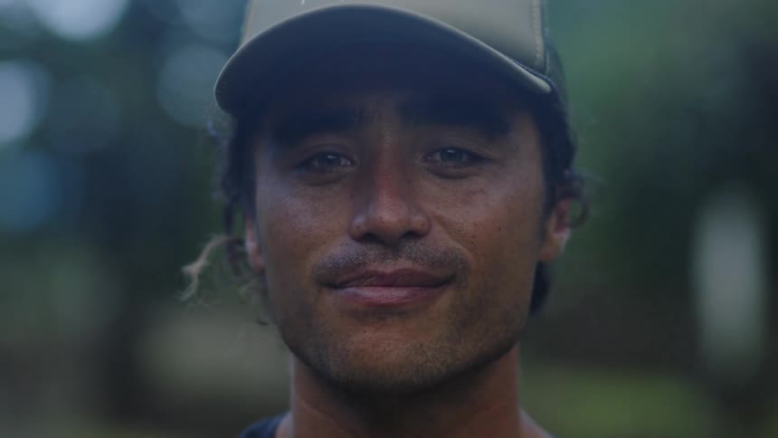 A person wearing a beige cap is standing outdoors with a background of trees and greenery