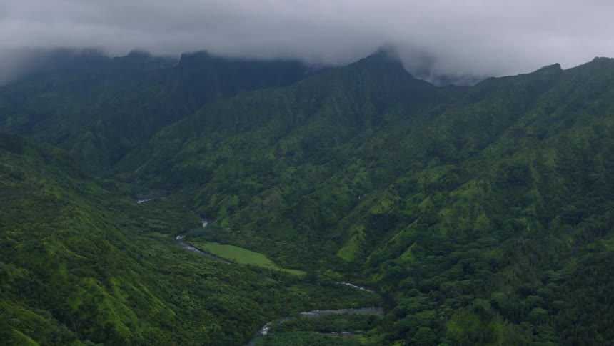 An aerial view shows a lush green valley and river surrounded by steep cloudcovered mountain peaks