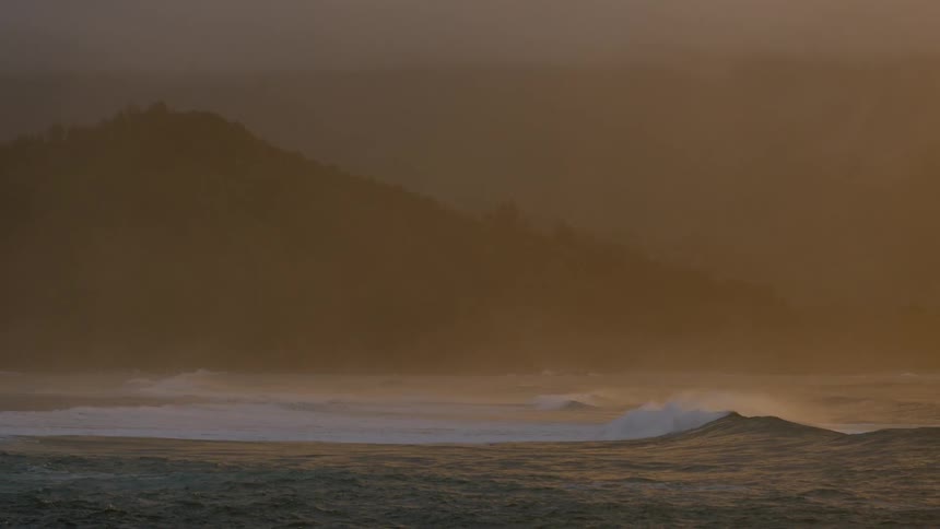 A wave curls and breaks along a hazy shoreline obscured by a misty yellowish atmosphere and the faint silhouette of a distant hill
