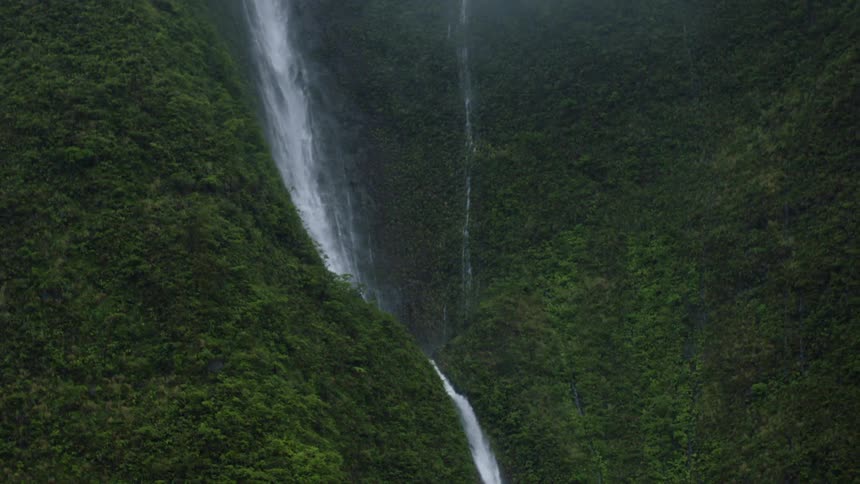 A tall narrow waterfall cascades down a lush green steep mountainside disappearing into the dense forest below