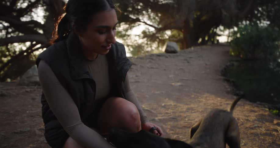A woman crouches next to her dog in a dirt path at sunset