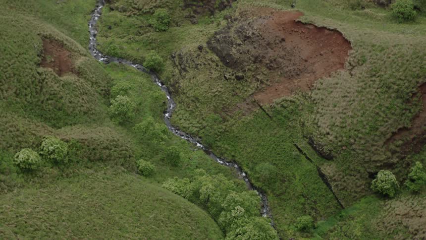 A thin stream meanders through a rugged grassy landscape dotted with patches of trees and rocky outcrops