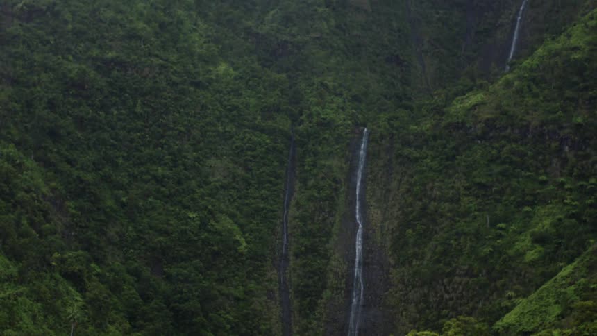 A camera pans up a steep lush mountain face revealing a tall thin waterfall cascading down the cliffs