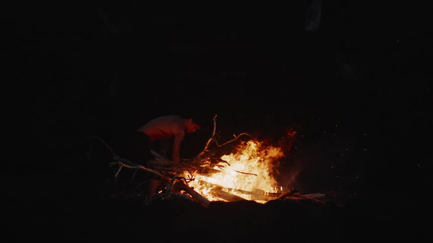 A man crouches near a small bonfire crackling with bright orange flames at night