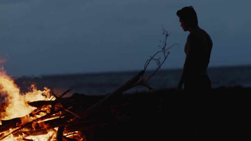 Silhouette of a man on the beach facing a bonfire in the sand