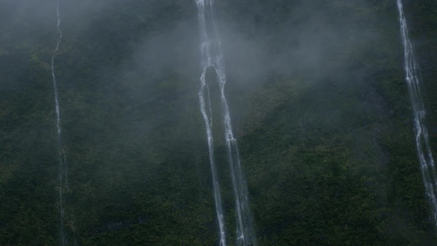 An aerial view shows thin waterfalls cascading down a lush green mountainside partially obscured by mist