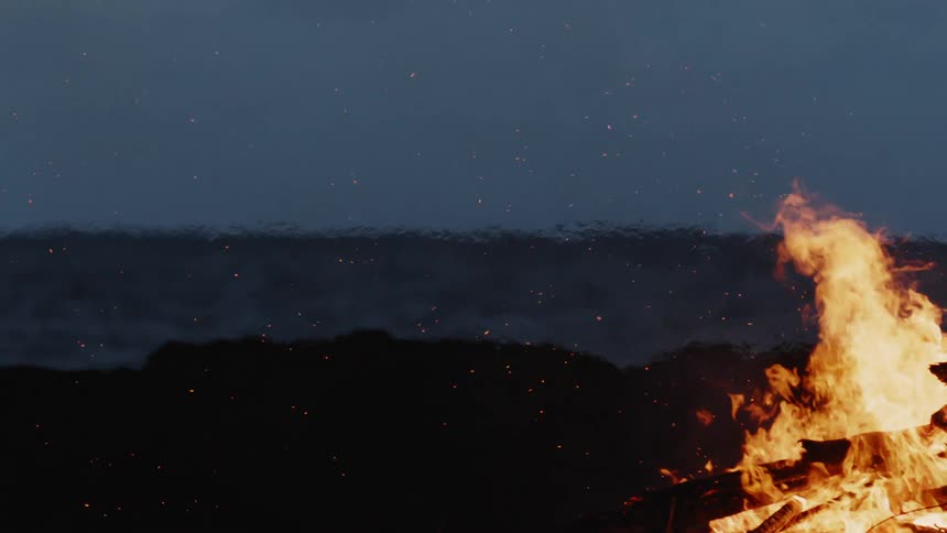 Pile of logs are burning with a dark mass of land in the background