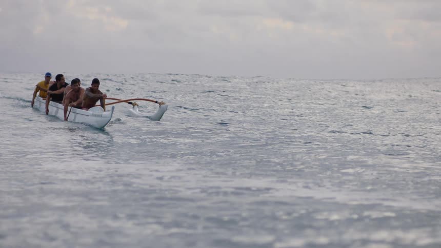 People are paddling a traditional outrigger canoe on a calm ocean under a cloudy sky