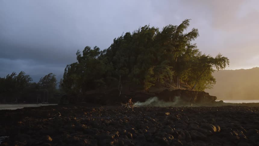 A rugged rocky shoreline with a dense tree covered island in the background bathed in warm golden sunlight while a lone person appears to be tending to a small fire with smoke rising into the air