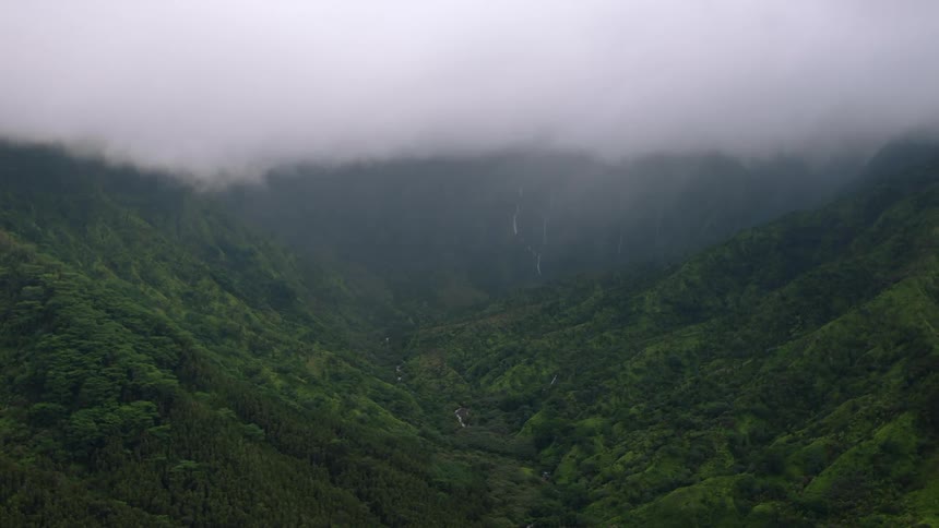A view down a lush green valley between cloudcovered mountains shows a river winding through the landscape