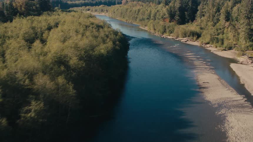 An aerial view shows a winding river course through a barren landscape revealing exposed earth and scattered debris alongside patches of remaining forest
