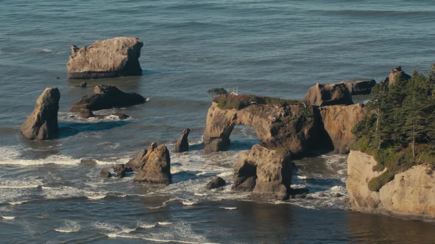 Jagged rock formations rise dramatically from the turbulent ocean waves with a natural arch and sea stacks visible