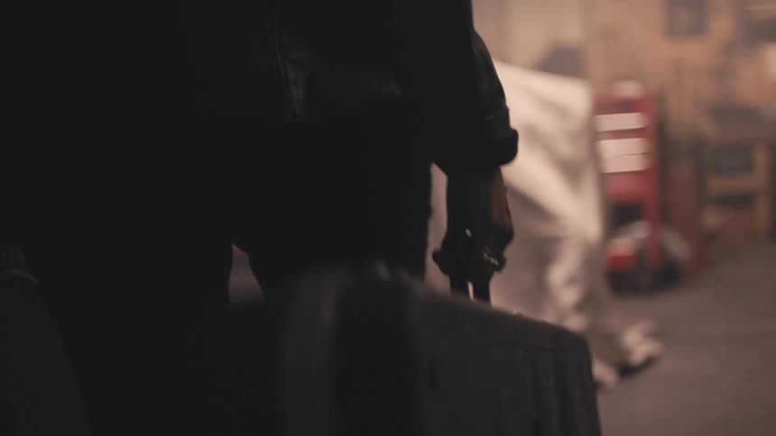 The lower half of a person in a leather jacket walking while carrying a guitar bag in one hand and a briefcase in the other in a dimly lit vintagestyle setting