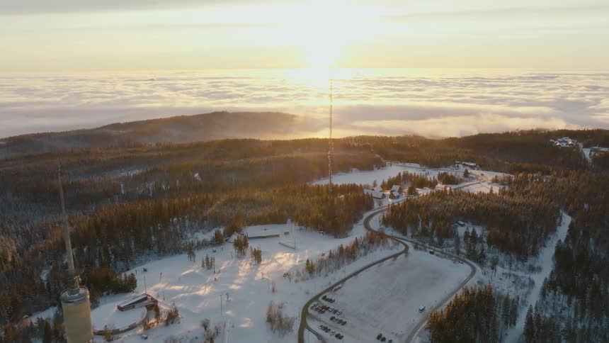Aerial view of a snowcovered mountainous area at dawn with a communication tower scattered buildings and a winding road between the trees