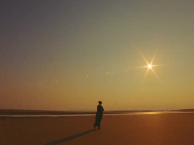 Silhouetted woman walks across the sandy terrain near the water under a bright sun