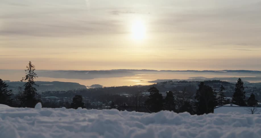 From a snowy hilltop the sun breaks through a cloudy sky casting a bright vertical ray over a distant hazy landscape with a winding river or road silhouetting trees in the foreground