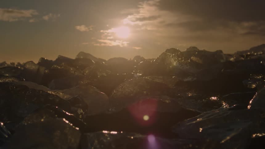Chunks of ice glisten on a beach under a bright sunlit sky with silhouetted figures in the distance