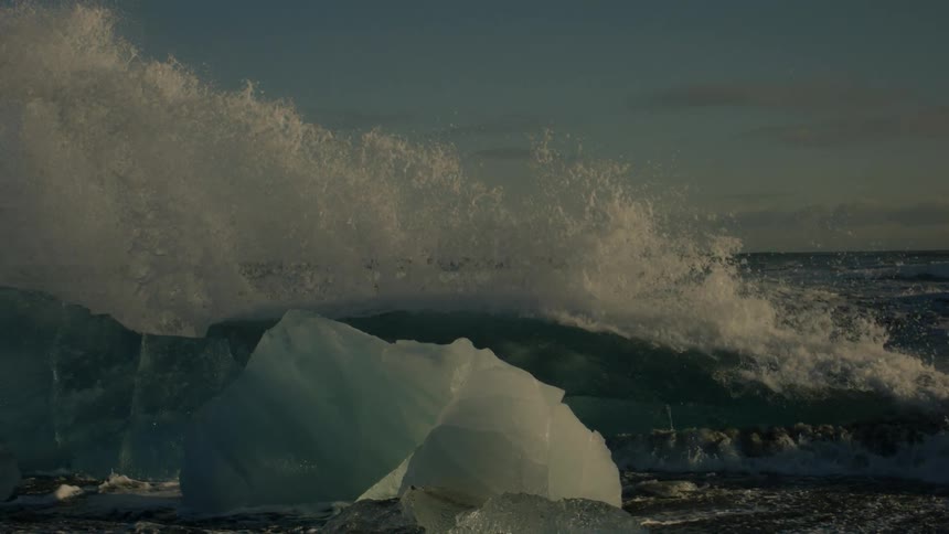 A large wave crashes over a rocky outcrop covered with large ice chunks sending white spray into the air